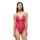 A model wearing a red patterned lace one-piece swimsuit with a deep V-neckline and thin shoulder straps, standing against a solid beige background.