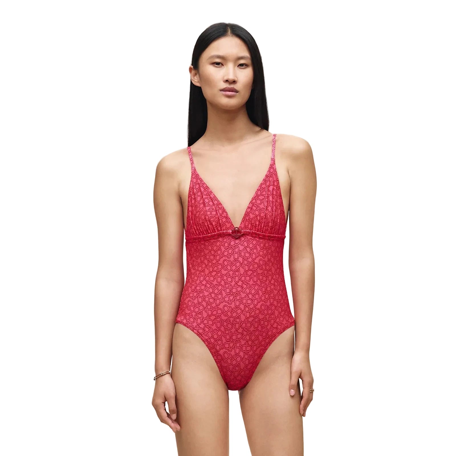 A model wearing a red patterned lace one-piece swimsuit with a deep V-neckline and thin shoulder straps, standing against a solid beige background.