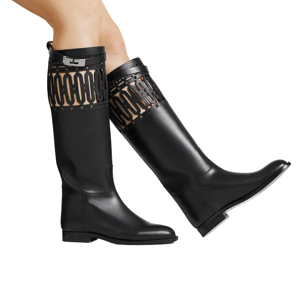 A pair of black leather knee-high boots with decorative cutout detailing near the top, shown on a model's legs against a light beige studio background.