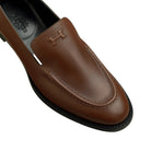 A close-up of a brown leather loafer by Hermès, showing the embossed H logo on the vamp and the brand name printed on the black insole.